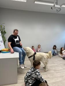 Na zdjęciu zgadują się uczestnicy zajęć. Na zdjęciu znajduje się pies. Uczestnicy siedzą w kręgu na poduszkach. W tle księgozbiór biblioteki.