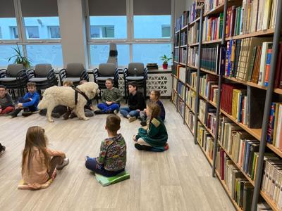 Na zdjęciu zgadują się uczestnicy zajęć. Na zdjęciu znajduje się pies. Uczestnicy siedzą w kręgu na poduszkach. W tle księgozbiór biblioteki.