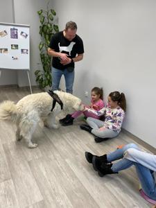 Na zdjęciu zgadują się uczestnicy zajęć. Na zdjęciu znajduje się pies. Uczestnicy siedzą w kręgu na poduszkach. W tle księgozbiór biblioteki.