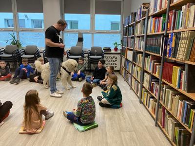 Na zdjęciu zgadują się uczestnicy zajęć. Na zdjęciu znajduje się pies. Uczestnicy siedzą w kręgu na poduszkach. W tle księgozbiór biblioteki.