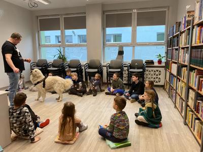 Na zdjęciu zgadują się uczestnicy zajęć. Na zdjęciu znajduje się pies. Uczestnicy siedzą w kręgu na poduszkach. W tle księgozbiór biblioteki.