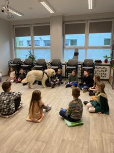 Na zdjęciu zgadują się uczestnicy zajęć. Na zdjęciu znajduje się pies. Uczestnicy siedzą w kręgu na poduszkach. W tle księgozbiór biblioteki.