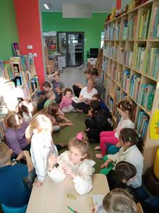 Zdjęcie przedstawia dzieci. Dzieci słuchają czytanej przez bibliotekarkę książki. Zdjęcie wykonano w bibliotece.