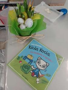 Zdjęcie przedstawia książkę i kwiaty w doniczce.  Zdjęcie wykonano na terenie biblioteki.