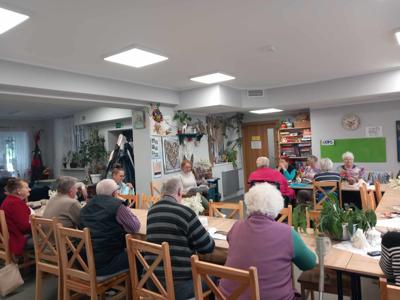 Zdjęcie przedstawia Seniorów. Seniorzy siedzą przy stołach, są zasłuchani w czytaną poezję