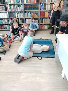 Na zdjęciu znajdują się strażacy i dzieci biorące udział w warsztatach pierwszej pomocy. Zdjęcie wykonano w bibliotece.