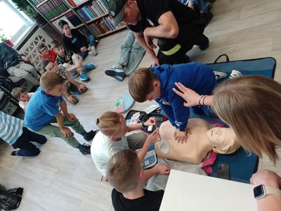 Na zdjęciu znajdują się strażacy i dzieci biorące udział w warsztatach pierwszej pomocy. Zdjęcie wykonano w bibliotece.