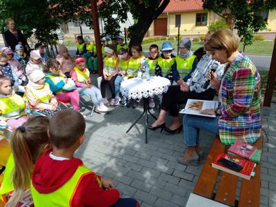 Zdjęcie przedstawia dzieci siedzące na krzesełkach i słuchające czytających lektorów.  Zdjęcie wykonano w altance na terenie biblioteki.