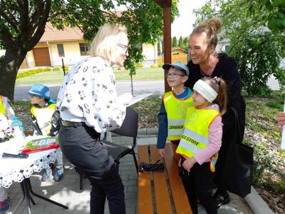Zdjęcie przedstawia dzieci siedzące na krzesełkach i słuchające czytających lektorów.  Zdjęcie wykonano w altance na terenie biblioteki.