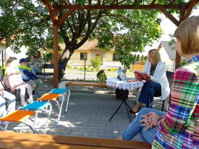 Zdjęcie przedstawia dzieci siedzące na krzesełkach i słuchające czytających lektorów.  Zdjęcie wykonano w altance na terenie biblioteki.