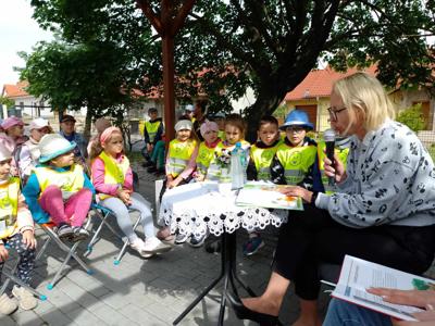 Zdjęcie przedstawia dzieci siedzące na krzesełkach i słuchające czytających lektorów.  Zdjęcie wykonano w altance na terenie biblioteki.