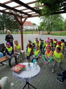 Zdjęcie przedstawia dzieci siedzące na krzesełkach i słuchające czytających lektorów.  Zdjęcie wykonano w altance na terenie biblioteki.