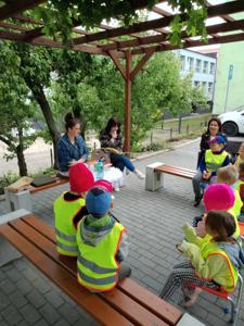Zdjęcie przedstawia dzieci siedzące na krzesełkach i słuchające czytających lektorów.  Zdjęcie wykonano w altance na terenie biblioteki.