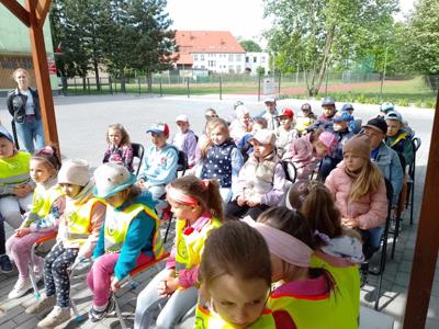 Zdjęcie przedstawia dzieci siedzące na krzesełkach i słuchające czytających lektorów.  Zdjęcie wykonano w altance na terenie biblioteki.