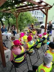 Zdjęcie przedstawia dzieci siedzące na krzesełkach i słuchające czytających lektorów.  Zdjęcie wykonano w altance na terenie biblioteki.
