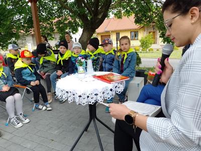 Zdjęcie przedstawia dzieci siedzące na krzesełkach i słuchające czytających lektorów.  Zdjęcie wykonano w altance na terenie biblioteki.