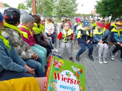 Zdjęcie przedstawia dzieci siedzące na krzesełkach i słuchające czytających lektorów.  Zdjęcie wykonano w altance na terenie biblioteki.
