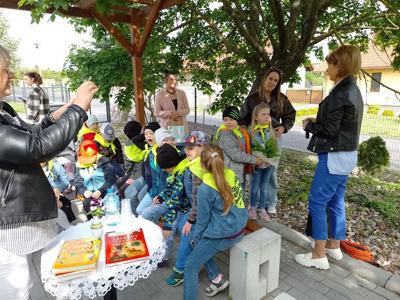 Zdjęcie przedstawia dzieci siedzące na krzesełkach i słuchające czytających lektorów.  Zdjęcie wykonano w altance na terenie biblioteki.