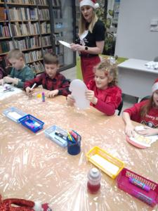 Na zdjęciu znajdują się dzieci siedzące przy stołach. Zdjęcie wykonano w bibliotece.