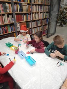 Na zdjęciu znajdują się dzieci siedzące przy stołach. Zdjęcie wykonano w bibliotece.