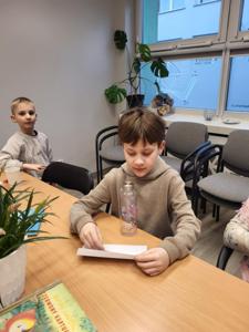 Na zdjęciu znajdują się uczestnicy zajęć. Uczestnicy siedzą. W tle książki.