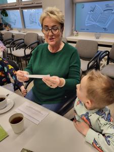 Na zdjęciu znajdują się uczestnicy zajęć. Uczestnicy siedzą. W tle książki.