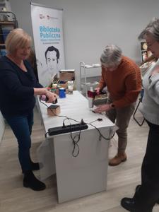 Na zdjęciu znajdują się Panie trzymające drewniane przedmioty. Zdjęcie wykonano w bibliotece.