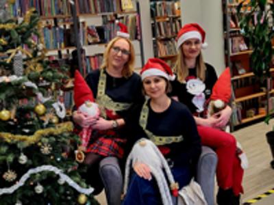 Na zdjęciu znajdują się 3 osoby trzymające w ręce skrzaty. W tle choinka i książki. Zdjęcie wykonano w bibliotece.