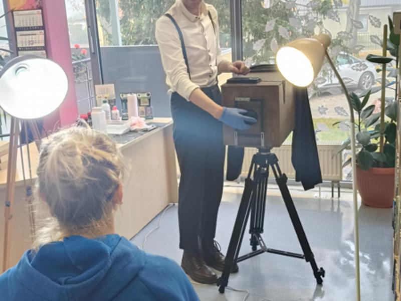 Fotograf odkrywa dekielek aparatu naprzeciw kobiety pozującej do zdjęcia.