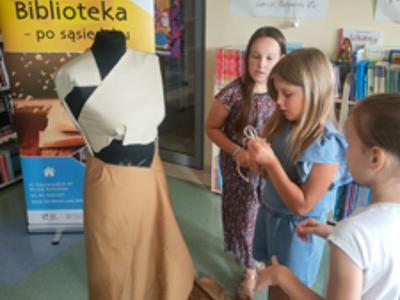 trzy dziewczynki przyglądają się manekinowi ubranemu w białą koszulkę i cielistą spódnicę
