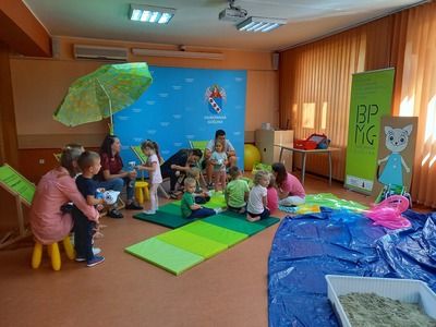 Grupa dzieci oraz ich rodziców siedzących na leżakach i matach słuchający kobiety, która czyta książkę
