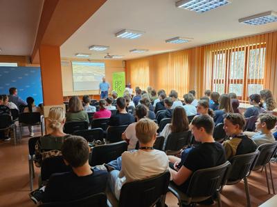 Grupa młodych ludzi siedzących na krzesłach i słuchajacych mówiącego mężyzczny