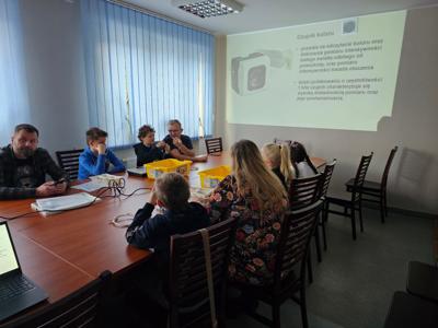 Grupa ludzi siedząca przy stole i patrząca na informacje wyświetlane na ścianie