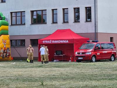 Na zdjęciu czerwony napis z napisem straż obok stoją strażacy w beżowych strojach