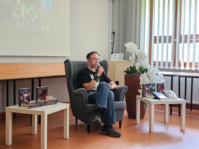 Na zjediu młody mężczyzna siedzący na szarym fotelu i trzymajacy mikrofon
