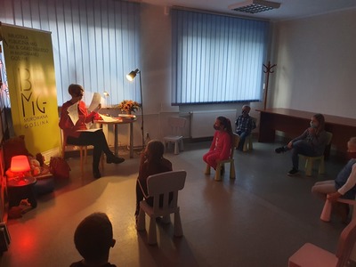 grupa dzieci siedzących na krzesłach i patrzących na kobietę w czerwonej bluzce