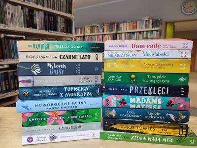 Książki, nowości wydawnicze dla dorosłych, leżące grzbietami na ladzie w tle regały biblioteczne.