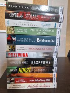 Książki, nowości wydawnicze dla dorosłych, leżące grzbietami na ladzie w tle regały biblioteczne.