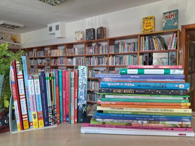 ksiązki stojące  z widocznymi grzbietami, obok ksiązki leżące z widocznymi grzbietami , w tle regały biblioteczne z ksiązkami