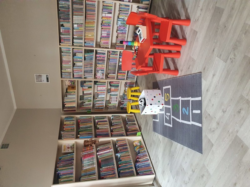 Na zdjęciu regały z książkami, czerwony stoliczek i krzesełka oraz zabawki