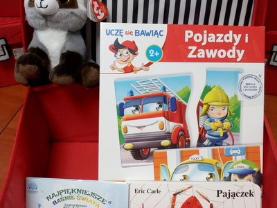 Na zdjęciu znajduje się czerwona otwarta walizeczka. W jej środku znajduje się gra edukacyjna, maskotka, audiobook i książka.