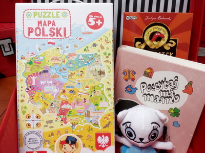 Na zdjęciu znajduje się czerwona otwarta walizeczka. W jej środku znajduje się gra edukacyjna, maskotka, audiobook i książka.