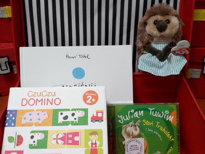 Na zdjęciu znajduje się czerwona otwarta walizeczka. W jej środku znajduje się gra edukacyjna, maskotka, audiobook i książka.