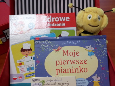 Na zdjęciu znajduje się czerwona otwarta walizeczka. W jej środku znajduje się gra edukacyjna, maskotka, audiobook i książka.
