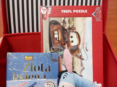 Na zdjęciu znajduje się czerwona otwarta walizeczka. W jej środku znajduje się gra edukacyjna, maskotka, audiobook i książka.