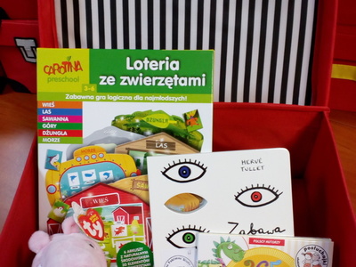Na zdjęciu znajduje się czerwona otwarta walizeczka. W jej środku znajduje się gra edukacyjna, maskotka, audiobook i książka.
