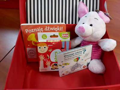 Na zdjęciu znajduje się czerwona otwarta walizeczka. W jej środku znajduje się gra edukacyjna, maskotka, audiobook i książka.