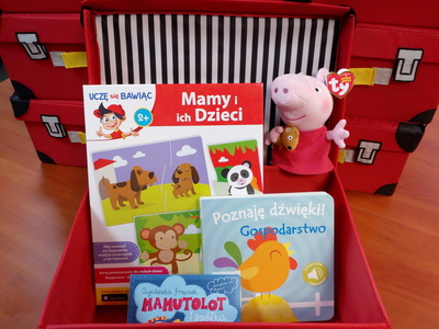 Na zdjęciu znajduje się czerwona otwarta walizeczka. W jej środku znajduje się gra edukacyjna, maskotka, audiobook i książka.