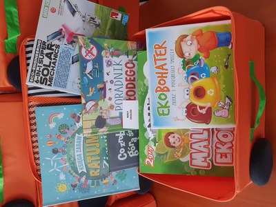 Na zdjęciu znajduje się pomarańczowa otwarta walizeczka. W jej środku znajduje się gra edukacyjna, książka, gadżet edukacyjny..