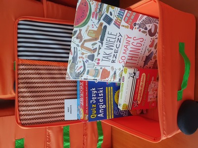 Na zdjęciu znajduje się pomarańczowa otwarta walizeczka. W jej środku znajduje się gra edukacyjna, książka, gadżet edukacyjny..